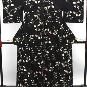 May include: A black kimono featuring a repeating pattern of delicate white and soft pink flowers with thin, winding green vines and stems. The garment has wide, rectangular sleeves and a long, straight silhouette, displayed on a black stand.