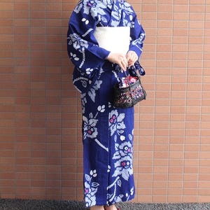 以下が含まれることがあります： 青色の浴衣を着た女性。白い花の模様、白い帯、赤いサンダル。小さな黒いハンドバッグには花の模様。浴衣は日本の伝統的な衣装です。