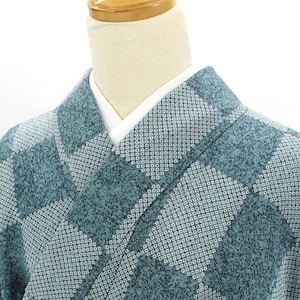 Puede incluir: Primer plano de un kimono color turquesa con un patrón geométrico. La tela tiene una apariencia texturizada con pequeños diseños cuadrados blancos. El kimono está drapeado sobre un maniquí, mostrando los detalles del cuello y los hombros. El fondo es blanco liso.