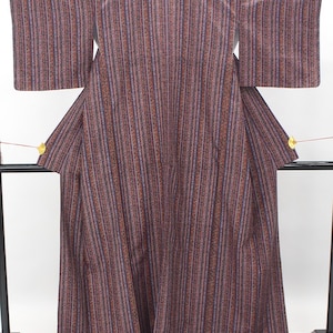 May include: A full-length kimono featuring a vertical striped pattern in shades of brown, dark blue, and tan. The garment has wide, rectangular sleeves and a structured collar, displayed on a black stand with small gold fan-shaped accents.