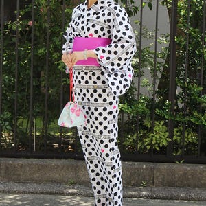 Puede incluir: Un yukata blanco con lunares negros grandes y un obi rosa. La prenda tradicional japonesa se usa con una bolsa con cordón a juego. El yukata presenta un diseño de corazón y se usa al aire libre.