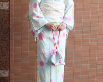May include: A light blue yukata with a floral pattern in pink, purple, and yellow. The traditional Japanese garment is tied with a white obi and includes a matching drawstring bag. The person is wearing sandals.