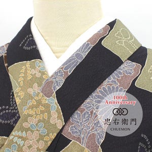 May include: A black kimono featuring a multi-colored floral and geometric pattern in shades of olive green, brown, and blue. The garment has a white inner collar and a circular logo overlay with the text 100th Anniversary and CHUEMON.