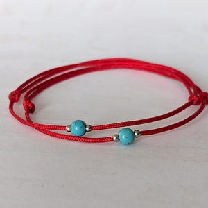 May include: Red string bracelet featuring two turquoise beads and silver accents. The bracelet is adjustable and knotted. The beads are a bright turquoise colour. The bracelet is on a white surface.