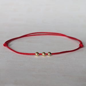 May include: A red string bracelet featuring three small, gold-coloured beads. The bracelet is knotted at each end. The background is a light, neutral colour.