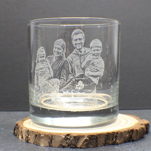 May include: A clear, engraved rocks glass featuring a family portrait. The glass sits on a round, natural wood coaster. The image is etched into the glass, showcasing a family of four. The bottom of the glass is filled with liquid.