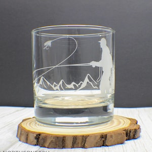 May include: A clear glass tumbler with a detailed etched design of a fly fisherman casting a line, with mountains in the background. The glass sits on a round, natural wood coaster. The design is in white.