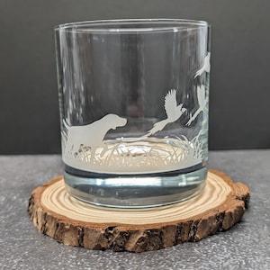 May include: A clear glass tumbler with etched artwork of a dog and birds in flight over tall grass. The glass sits on a round, natural wood coaster with a bark edge. The design is suitable for a hunting or outdoors theme.