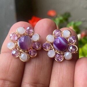 May include: A pair of handcrafted floral stud earrings. Each earring features a central purple gemstone surrounded by smaller pink and white beads, all set in a gold-toned wire frame. The earrings are designed to resemble a flower.