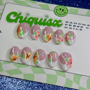 May include: Handmade press-on nails with a floral design. The nails feature a light pink base with a white tip and a colorful flower design in orange, green, and pink. The brand name "Chiquisoxx" is visible on the packaging.