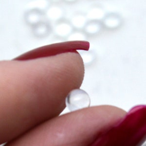 May include: A clear, round, plastic bead held between two fingers with red nail polish.