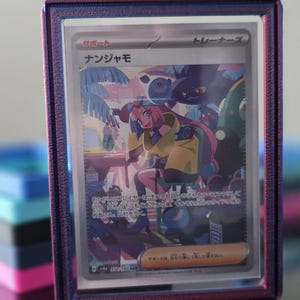 May include: A framed trading card featuring a colorful illustration of a character in a yellow coat and pink hair. The card has Japanese text and is encased in a blue and purple frame, resting on a reflective surface.