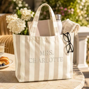 May include: A beige and white vertical striped tote bag featuring the embroidered text MRS. CHARLOTTE on the front. The bag contains a bouquet of white flowers, a notebook, a silver water bottle, a folded cloth, and black-rimmed eyeglasses.