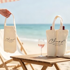 May include: Off-white canvas wine tote bags with black script that reads "Cheers" and a heart. The bags also include names and a date. One bag holds a single bottle, and the other holds two bottles. A glass of rose wine sits on a wooden table.