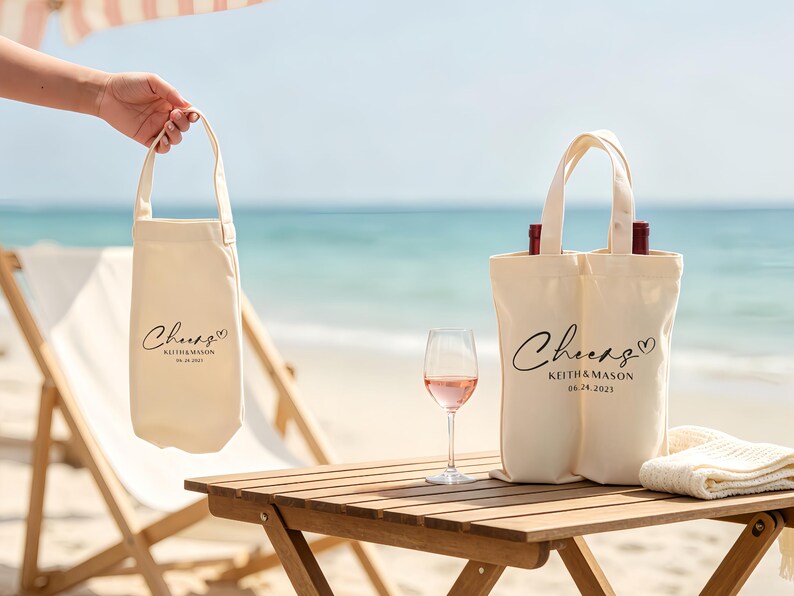 May include: Off-white canvas wine tote bags with black script that reads "Cheers" and a heart. The bags also include names and a date. One bag holds a single bottle, and the other holds two bottles. A glass of rose wine sits on a wooden table.