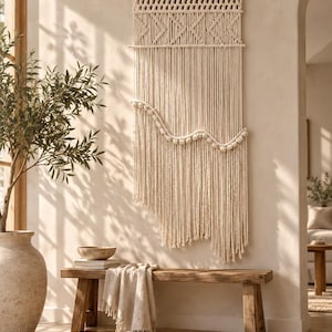 May include: A large off-white macrame wall hanging suspended from a wooden dowel. The piece features intricate knotted patterns at the top, a horizontal wavy section adorned with small round knots, and long, flowing fringe extending to the bottom.
