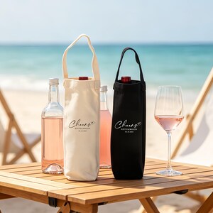 May include: A beach scene features a wooden table with wine bottles and glasses. Two wine bottle tote bags, one cream and one black, are visible. The cream bag has a white handle. The text "Cheers" is printed on the bags. A glass of rose wine is on the table.