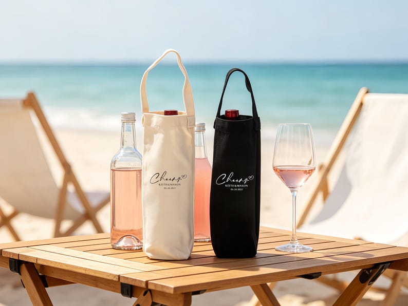 May include: A beach scene features a wooden table with wine bottles and glasses. Two wine bottle tote bags, one cream and one black, are visible. The cream bag has a white handle. The text "Cheers" is printed on the bags. A glass of rose wine is on the table.