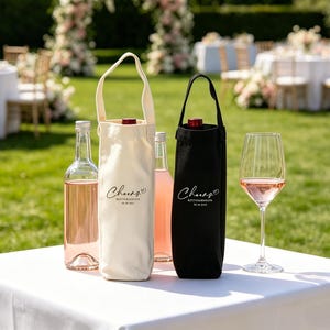 May include: A white table displays wine bottles, wine totes, and a wine glass. The bottles contain pink wine. One tote is black, the other is off-white, both with "Cheers" text. A wedding reception is in the background.