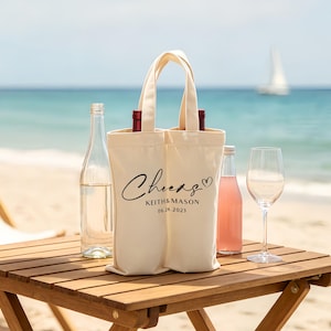 May include: A cream-colored wine tote bag with the word "Cheers" and the names "Keith & Mason" and the date "06.24.2021" printed on it. The bag holds two bottles of wine. A clear wine bottle, a pink bottle, and a wine glass are on a wooden table.