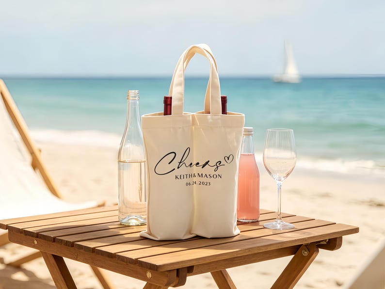 May include: A cream-colored wine tote bag with the word "Cheers" and the names "Keith & Mason" and the date "06.24.2021" printed on it. The bag holds two bottles of wine. A clear wine bottle, a pink bottle, and a wine glass are on a wooden table.