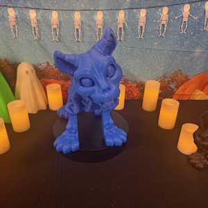 May include: A blue cat-like Halloween ornament with a skeletal face and paws. The cat is surrounded by lit candles, ghost figures, and a pumpkin. A string of skeleton lights hangs in the background.