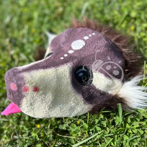 May include: A plush animal head mask featuring dark brown and cream fur, a prominent black eye, a bright pink tongue protruding from the mouth, and a series of white circular markings along the forehead, with a white tuft of fur at the ear.