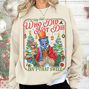 May include: Beige sweatshirt featuring a Christmas-themed graphic with Gonzo and Rizzo the Rat. The graphic includes text that reads "Who Did Not Die" and "Isn't That Swell". The design includes Christmas trees, ornaments, and gift boxes.