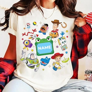 May include: Cream-colored t-shirt featuring colorful cartoon characters and the word "NAME" inside a green frog-shaped frame. The design includes a rocket, stars, and various toy-themed illustrations.