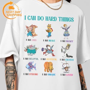 May include: White t-shirt with the text "I CAN DO HARD THINGS" and illustrations of cartoon characters. Below the illustrations are the phrases "I AM KIND", "I AM BRAVE", "I AM SMART", "I AM HELPFUL", "I AM GRATEFUL", "I AM ENOUGH", "I AM STRONG", "I AM UNIQUE", and "I AM LOVED".