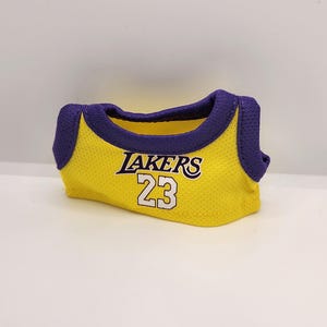 Pu&ograve; includere: Una maglia gialla in rete con finiture viola. La maglia presenta la scritta "LAKERS" in viola e bianco, insieme al numero "23" in bianco. La maglia &egrave; piegata e sembra essere realizzata in un tessuto traspirante.