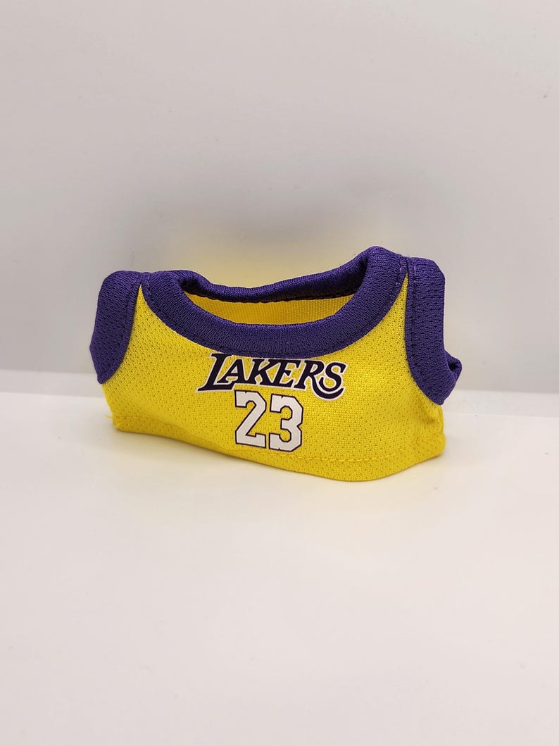 Pu&ograve; includere: Una maglia gialla in rete con finiture viola. La maglia presenta la scritta "LAKERS" in viola e bianco, insieme al numero "23" in bianco. La maglia &egrave; piegata e sembra essere realizzata in un tessuto traspirante.