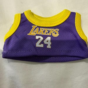 Pu&ograve; includere: Una maglia da basket in miniatura viola e gialla. La maglia presenta la scritta "Lakers" in lettere gialle e il numero "24" in bianco. Ha colletto e finiture gialle.