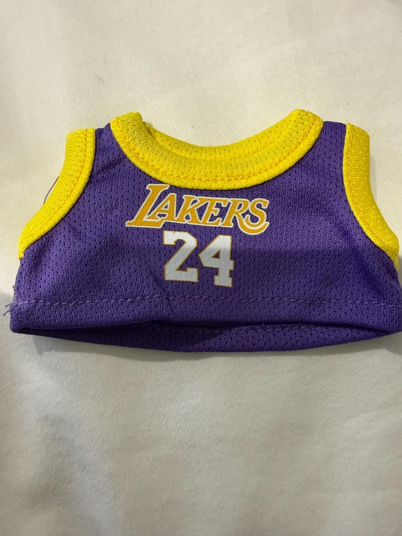 Pu&ograve; includere: Una maglia da basket in miniatura viola e gialla. La maglia presenta la scritta "Lakers" in lettere gialle e il numero "24" in bianco. Ha colletto e finiture gialle.
