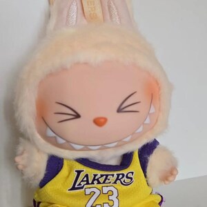 Pu&ograve; includere: Un peluche raffigurante un personaggio dei cartoni animati sorridente con orecchie da coniglio, che indossa una maglia gialla e viola dei Lakers con il numero 23. Il giocattolo ha un cappuccio di pelliccia color crema e un portachiavi.
