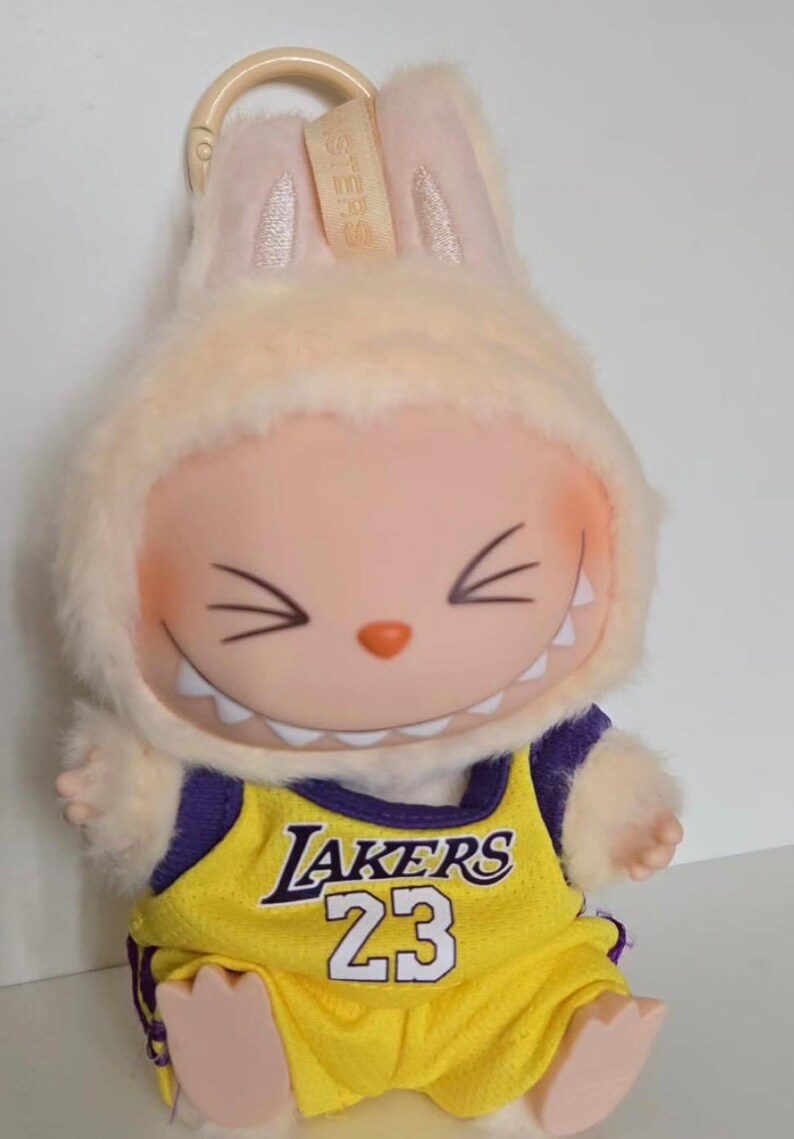 Pu&ograve; includere: Un peluche raffigurante un personaggio dei cartoni animati sorridente con orecchie da coniglio, che indossa una maglia gialla e viola dei Lakers con il numero 23. Il giocattolo ha un cappuccio di pelliccia color crema e un portachiavi.