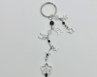 May include: Silver keychain with a dangling design featuring musical notes, stars, and beads. The keychain has a large ring at the top and a flower-shaped charm at the bottom. The beads are black and white.