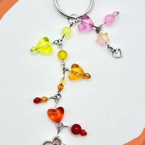 May include: A colorful keychain with a silver ring. The charm features a series of beads in yellow, orange, pink, and red, including heart and star shapes. The keychain is a decorative accessory.