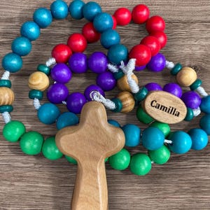 May include: A vibrant rosary featuring wooden beads in blue, green, purple, and red. It includes a wooden cross and an oval tag with the name "Camilla". The rosary is presented on a wooden background.