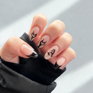 May include: A hand displaying long, almond-shaped nails. The nails have a clear base with black flame designs and black French tips. The hand is set against a light grey backdrop, with a black sleeve visible.