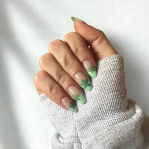 May include: A hand with oval nails, each adorned with a light green star design on a clear base. The tips are painted light green. The hand is wearing a grey sweatshirt.