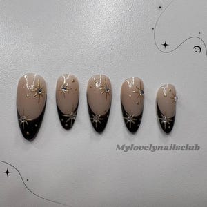 Pode incluir: Conjunto de cinco unhas postiças em formato amêndoa. Cada unha apresenta uma ponta francesa preta, uma base nude e detalhes dourados de estrelas e strass. O texto "Mylovelynailsclub" é visível.