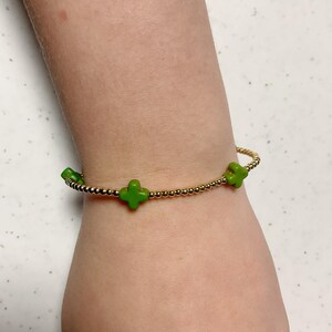 May include: Gold beaded bracelet featuring three bright green clover charms. The bracelet is worn on a wrist, highlighting the delicate design and vibrant colour of the charms. A stylish accessory.