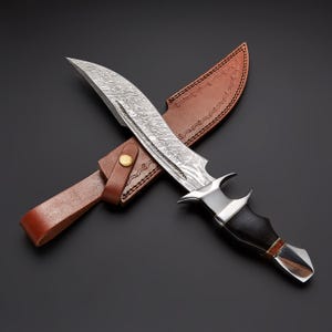 Può includere: Un grande coltello da caccia con una lama a motivi e un manico in legno scuro. Il coltello è abbinato a un fodero in pelle marrone. La lama è argentata e il manico è nero. Il fodero è marrone con cuciture decorative.