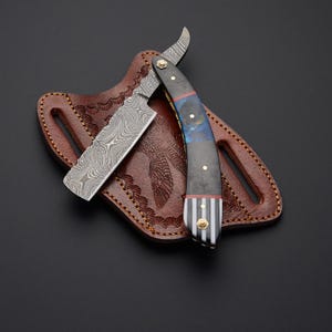 May include: A straight razor with a patterned blade and a decorative handle featuring blue and red accents, resting on a brown leather sheath. The sheath has an embossed design.