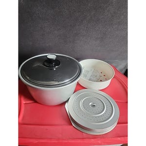 Puede incluir: Una arrocera blanca y plateada con tapa negra y asa blanca. La tapa está cerrada, y hay una cesta de vapor blanca y una placa perforada plateada al lado. La arrocera está sobre una superficie roja.