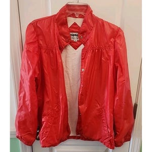 May include: A bright red vintage jacket with a high collar and snap closures. The jacket has a slightly shiny finish and two front pockets. The interior lining is a light beige color. The label reads "Windy Wear".
