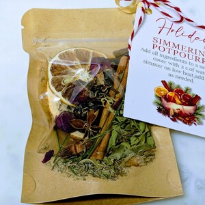 May include: A clear plastic pouch filled with dried herbs, spices, and a dried lemon slice. The pouch is sealed inside a brown paper bag. A tag with red and white twine reads "Holiday Simmering Potpourri".