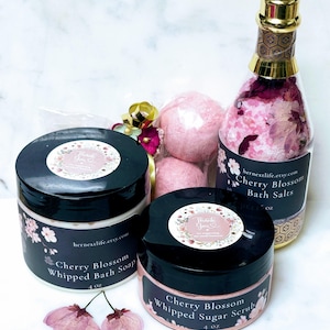May include: A collection of cherry blossom-scented bath products. Includes a bottle of bath salts, two jars of whipped soap and scrub, and three bath bombs. The products are in black jars with floral labels and a clear bottle. The bottle is shaped like a champagne bottle.