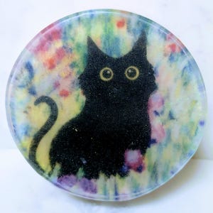 May include: Round, decorative cat art piece. The black cat has large yellow eyes and a long tail. The background is a colorful mix of red, pink, yellow, and blue. The cat is centered on the round piece.
